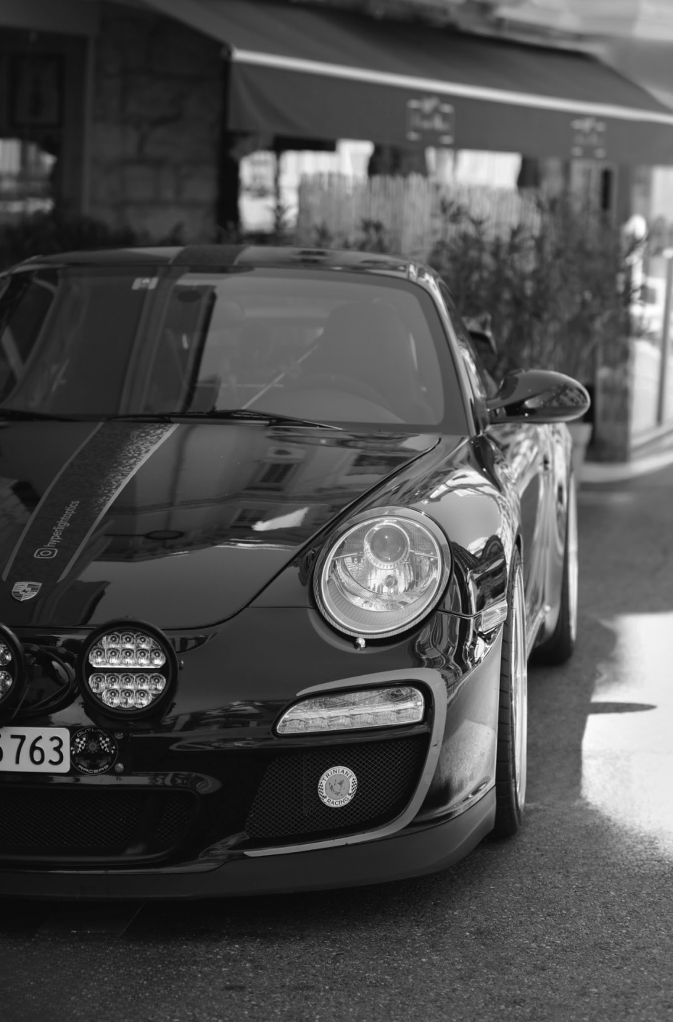 Porsche noire photographiée de face dans une rue bordée de terrasses
